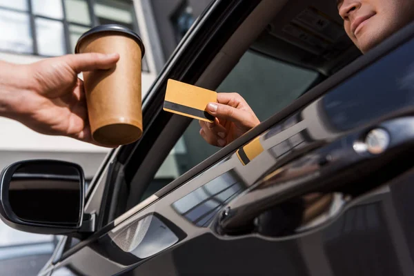 Vista cortada do homem segurando cartão de crédito enquanto sentado no carro perto do vendedor com café para ir em primeiro plano desfocado — Fotografia de Stock