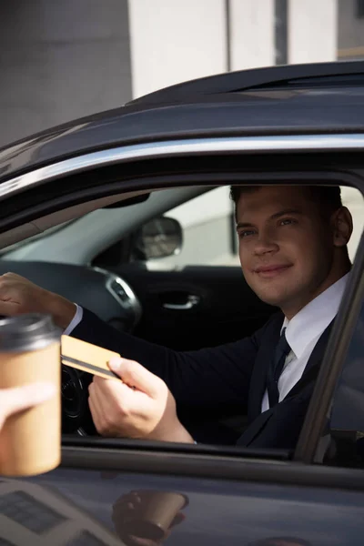 Sonriente hombre de negocios con tarjeta de crédito en auto cerca del vendedor con café para llevar en primer plano borrosa - foto de stock