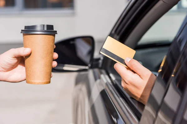 Vista recortada del vendedor sosteniendo café para acercarse al hombre con tarjeta de crédito en auto - foto de stock