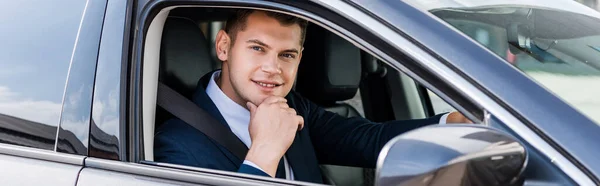 Uomo d'affari sorridente con mano vicino al mento guardando la fotocamera in auto, banner — Foto stock