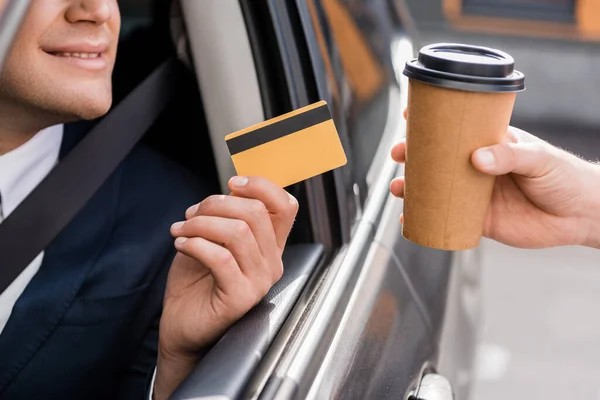 Vista recortada de hombre de negocios sonriente en el coche con tarjeta de crédito cerca del vendedor con café para llevar - foto de stock