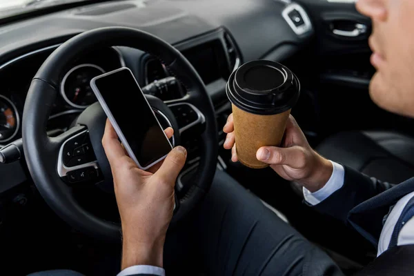 Vista recortada de hombre de negocios sosteniendo teléfono inteligente con pantalla en blanco y café para llevar en auto - foto de stock