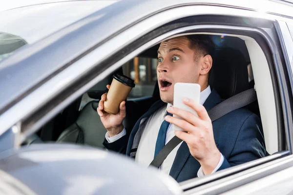 Uomo d'affari impaurito che tiene il caffè per andare e smartphone in auto in primo piano sfocato — Foto stock
