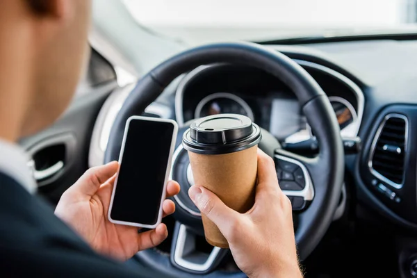 Обрезанный вид человека, держащего смартфон с чистым экраном и кофе на вынос в машине на размытом фоне — стоковое фото