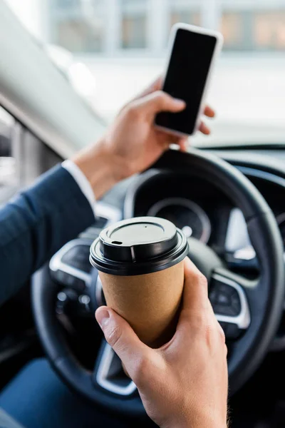 Vista recortada del hombre de negocios sosteniendo café para llevar y teléfono inteligente sobre fondo borroso en el coche - foto de stock