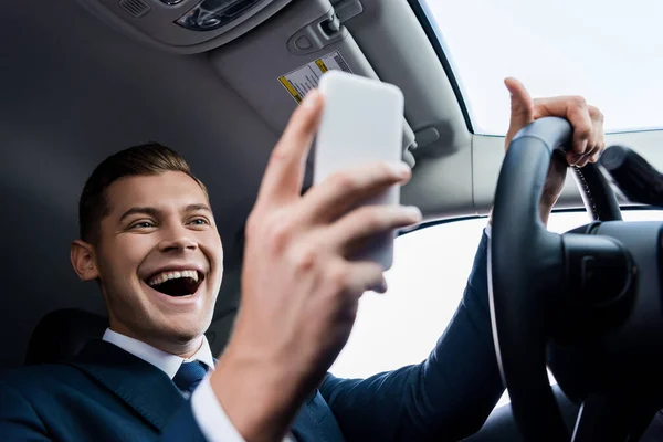 Low angle view of cheerful businessman driving auto and using smartphone on blurred foreground — Stock Photo