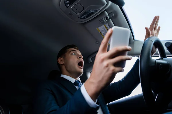 Vista de ángulo bajo del hombre de negocios asustado sosteniendo el teléfono inteligente en primer plano borroso en el coche - foto de stock