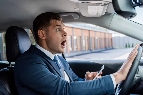 Empresario sorprendido sosteniendo teléfono inteligente y presionando la bocina del coche - foto de stock