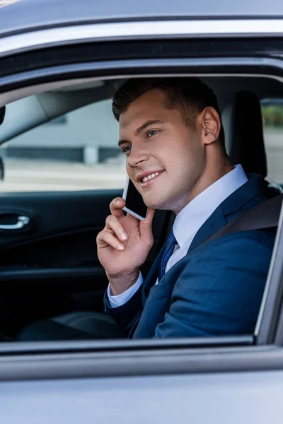 Hombre de negocios sonriente en ropa formal hablando en el teléfono inteligente en auto - foto de stock