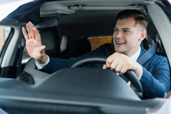 Cheerful businessman waving hand while driving car on blurred foreground — Stock Photo