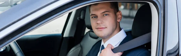 Jeune homme d'affaires regardant la caméra tout en tenant la ceinture de sécurité dans la voiture sur le premier plan flou, bannière — Photo de stock