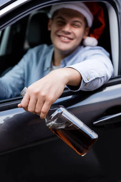 Drunk man in santa hat smiling and holding bottle of whiskey in car on blurred background — Stock Photo