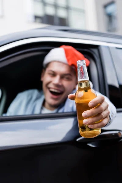 Borracho, hombre riendo en el sombrero de santa sosteniendo la botella de whisky en la mano extendida mientras está sentado en el coche, borrosa primer plano - foto de stock