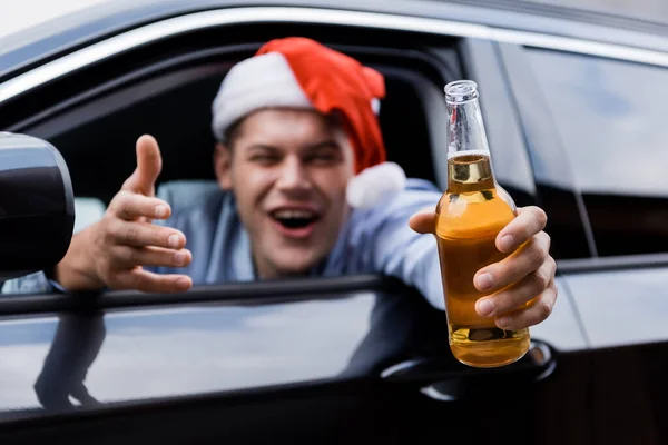 Drunk man in santa hat looking at camera while sitting in car and holding bottle of whiskey in outstretched hand — Stock Photo