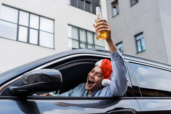 Excited, drunk man in santa hat holding bottle of alcohol in raised hand while sitting in car — Stock Photo