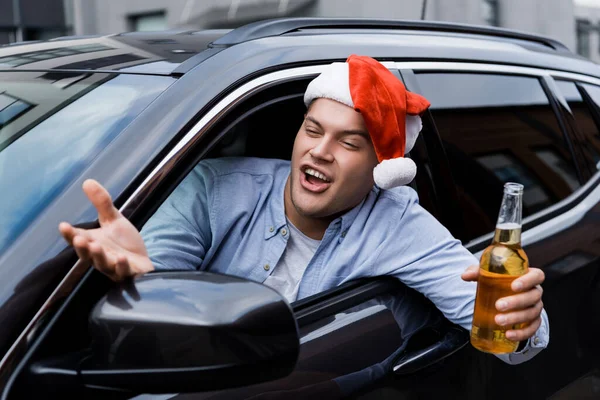 Excitado, hombre borracho en sombrero de santa, con frasco de alcohol, gesto mientras mira por la ventana del coche - foto de stock