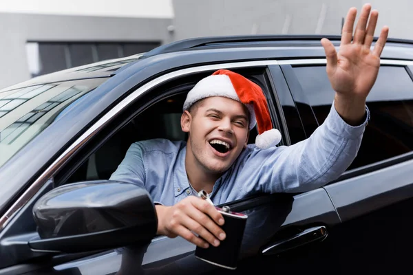 Betrunkener, aufgeregter Mann mit Weihnachtsmannmütze, winkt mit der Hand und hält eine Flasche Alkohol in der Hand, während er aus dem Autofenster schaut — Stockfoto