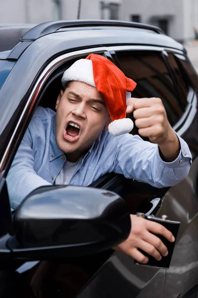 Aggressivo, uomo ubriaco in cappello di Babbo Natale, con fiaschetta di alcol, mostrando pugno serrato mentre si guarda fuori dal finestrino dell'auto, primo piano sfocato — Foto stock