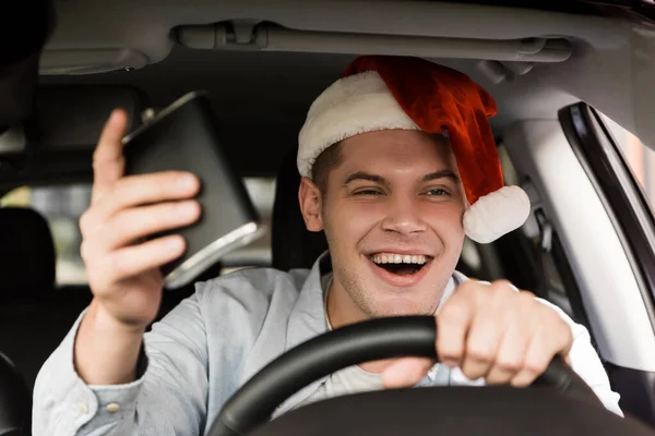 Drunk, cheerful man in santa hat holding flask with alcohol while driving car, blurred foreground — Stock Photo