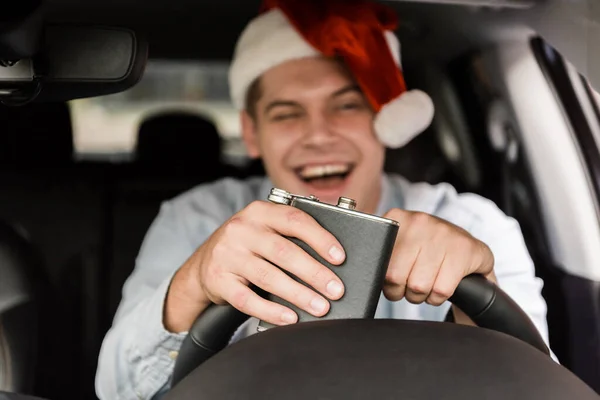 Laughing, drunk man in santa hat holding flask with alcohol while driving car on blurred background — Stock Photo