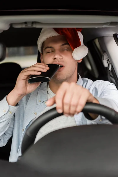 Drunk man in santa hat drinking from flask with closed eyes while driving car on blurred foreground — Stock Photo