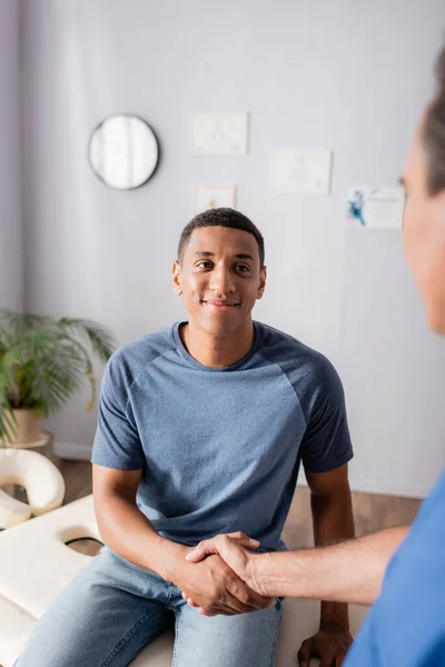 Happy african american patient shaking hands with chiropractor on blurred foreground — Stock Photo