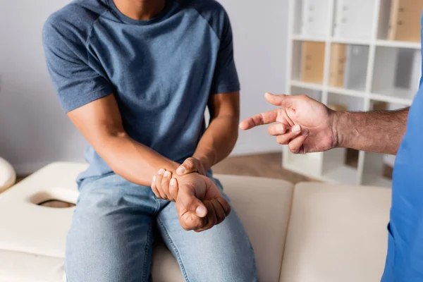 Vista recortada del quiropráctico apuntando con el dedo al brazo lesionado del paciente afroamericano - foto de stock