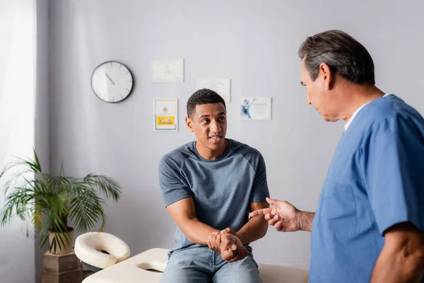 Quiropráctico señalando con el dedo al brazo lesionado del paciente afroamericano - foto de stock