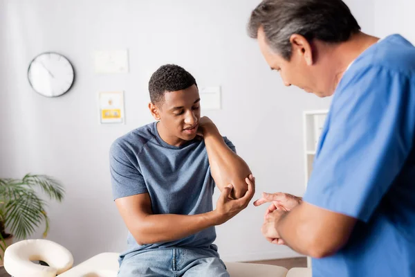 Hombre afroamericano lesionado mostrando codo a doctor en clínica - foto de stock