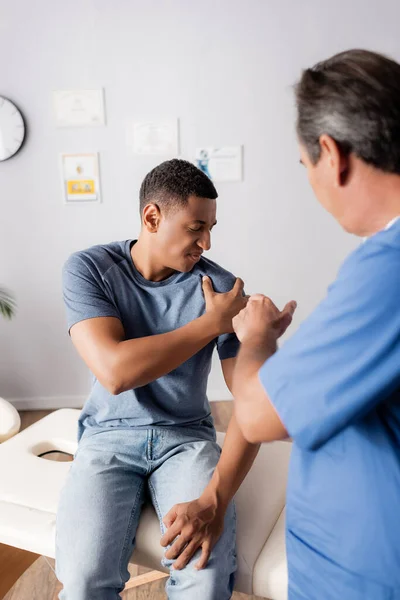 Doctor borroso señalando brazo lesionado de paciente afroamericano - foto de stock