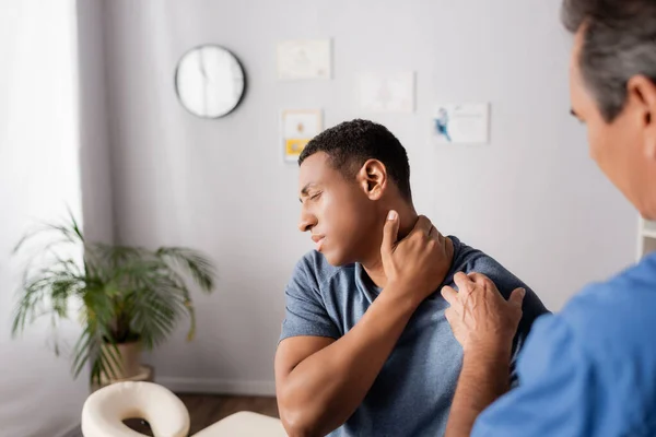 Herido africano americano hombre tocando cuello cerca doctor en borrosa primer plano - foto de stock