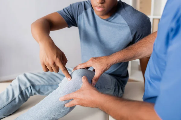 Обрезанный вид африканского американского пациента, указывающего пальцем на колено рядом с доктором — стоковое фото