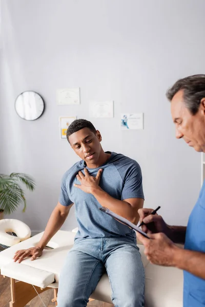 Doctor writing prescription on clipboard near african american patient in clinic — Stock Photo