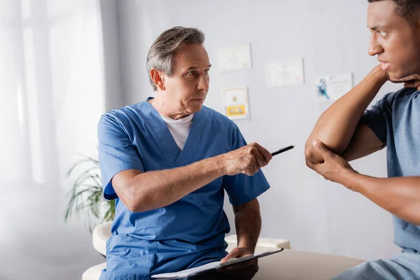 Medico che tiene la penna mentre indica il gomito ferito del paziente afroamericano in clinica — Foto stock
