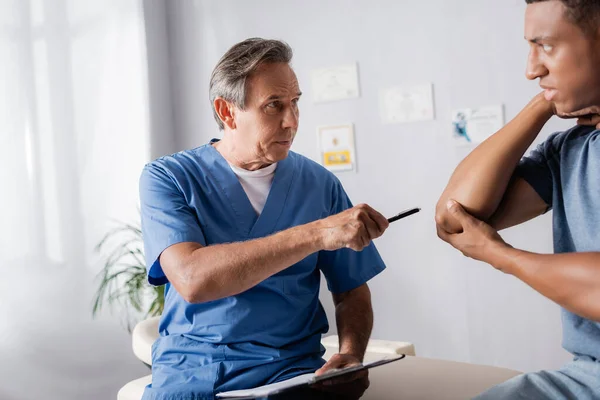 Médico sosteniendo la pluma mientras señala el codo lesionado del paciente afroamericano en la clínica - foto de stock