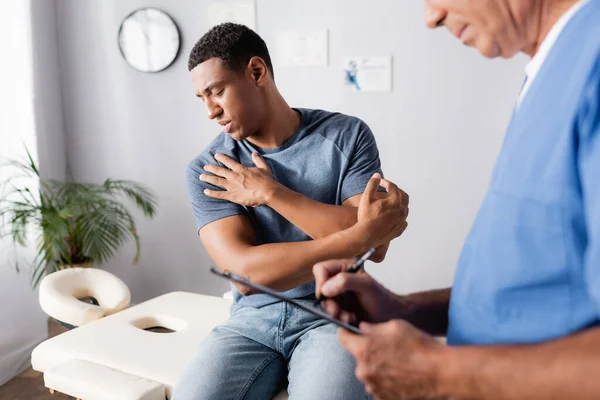 Verletzter afrikanisch-amerikanischer Mann blickt auf Arm in der Nähe des Arztes und schreibt Rezept auf verschwommenem Vordergrund — Stockfoto