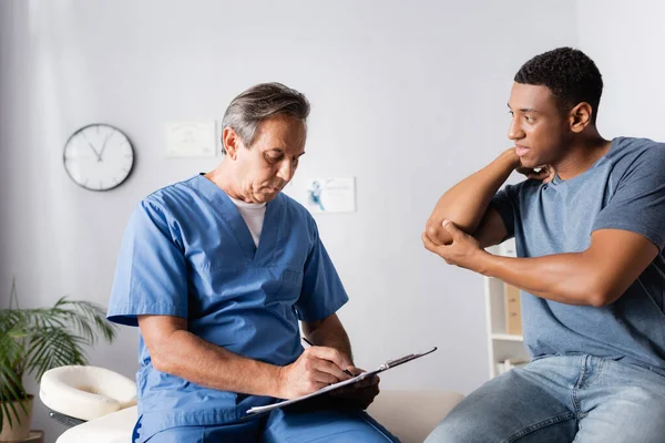 Mature doctor writing prescription near injured african american man — Stock Photo