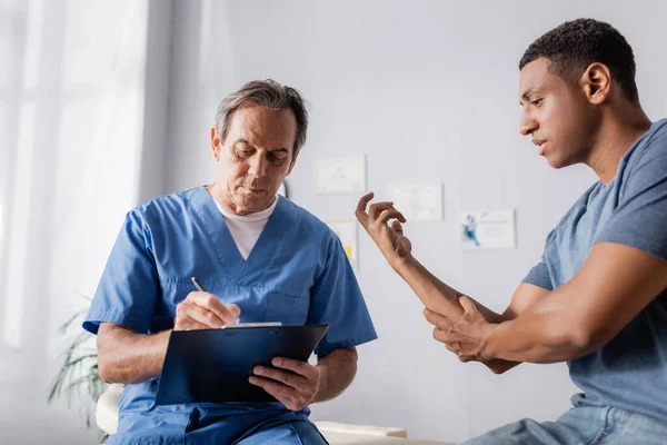 Doctor maduro escribiendo prescripción en portapapeles cerca del hombre afroamericano lesionado - foto de stock