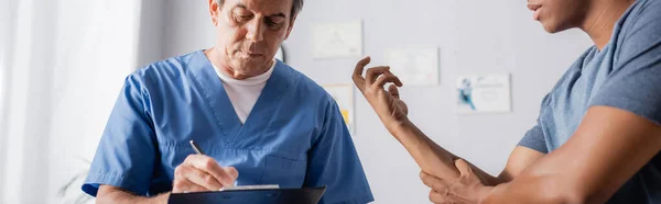 Mature doctor writing prescription on clipboard near injured african american man, banner — Stock Photo