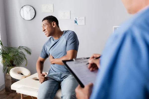 African american patient suffering from pain near doctor writing prescription on blurred foreground — Stock Photo