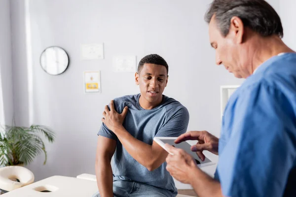 Paciente afroamericano que sufre de dolor cerca del médico usando tableta digital - foto de stock