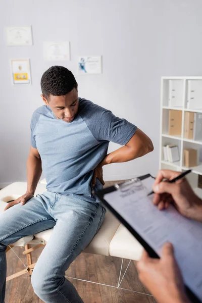 Young african american patient suffering from pain in back near doctor writing prescription on blurred foreground — Stock Photo