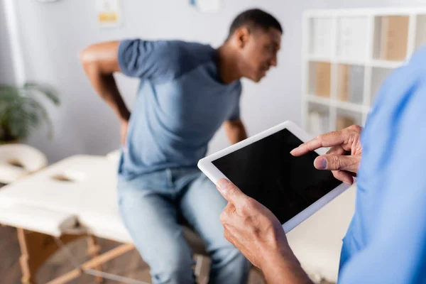 Mature doctor pointing with finger at digital tablet near african american patient suffering from pain on blurred background — Stock Photo