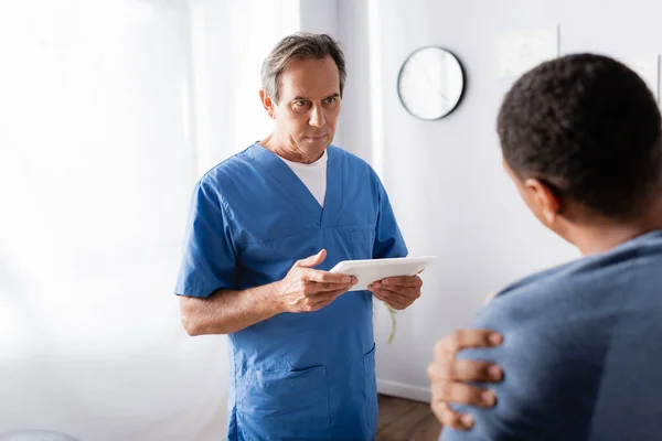 Medico maturo in possesso di tablet digitale vicino afro-americano paziente in primo piano offuscata — Foto stock