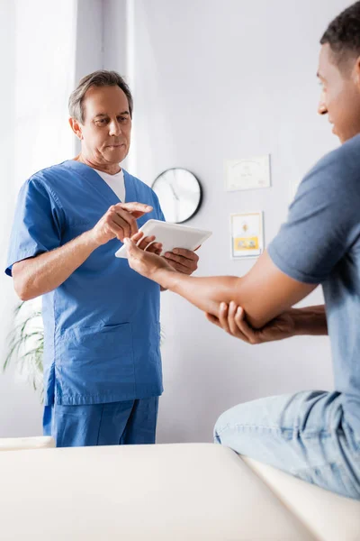 Mature doctor holding digital tablet and pointing with finger at african american patient on blurred foreground — Stock Photo