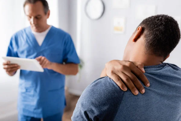 Uomo afroamericano ferito toccando collo vicino al medico con tablet digitale su sfondo sfocato — Foto stock