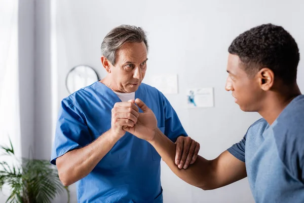 Therapist working with injured arm of african american man in clinic — Stock Photo