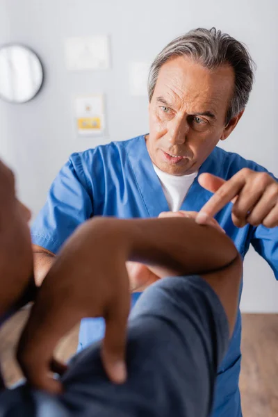 Terapeuta señalando con el dedo a la mano lesionada del paciente afroamericano en primer plano borroso - foto de stock