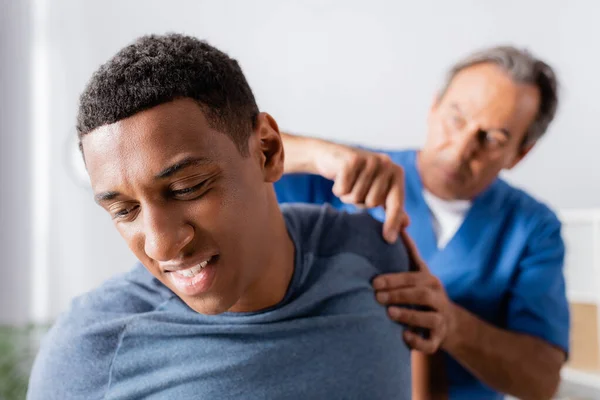 Afrikanisch-amerikanischer Mann leidet unter Schmerzen in der Nähe von Masseur auf verschwommenem Hintergrund — Stockfoto