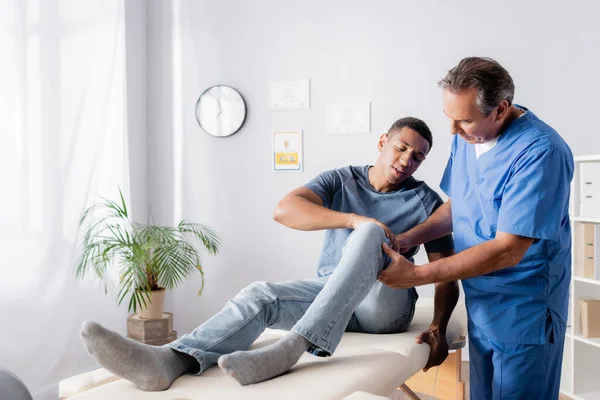 Älterer Chiropraktiker berührt lädiertes Knie eines afrikanisch-amerikanischen Patienten — Stockfoto